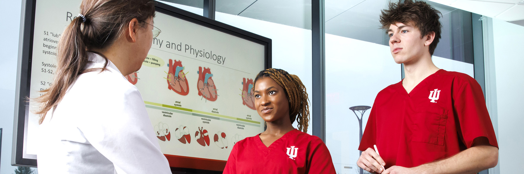 A nursing faculty member stands with two nursing students in red scrubs near a television with information on the screen.
