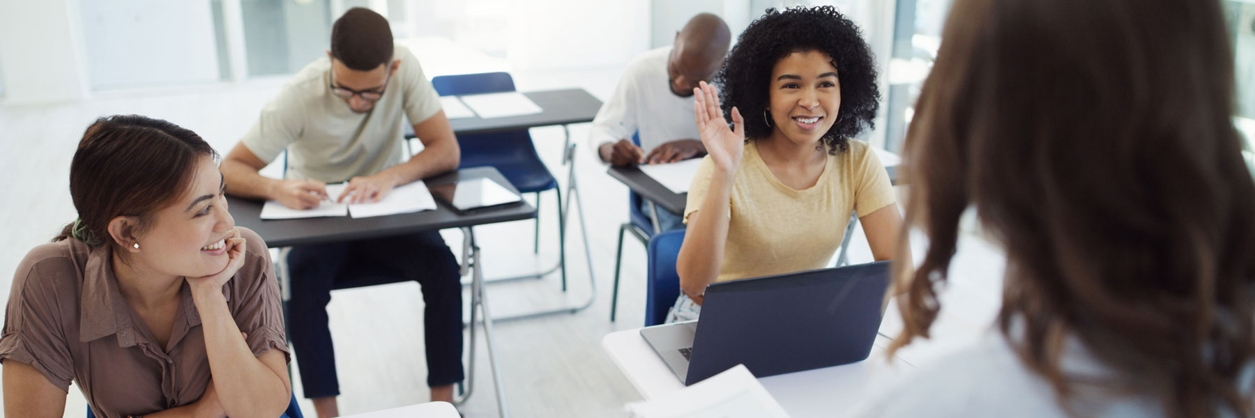 A student raises their hand in a classroom while others write or listen