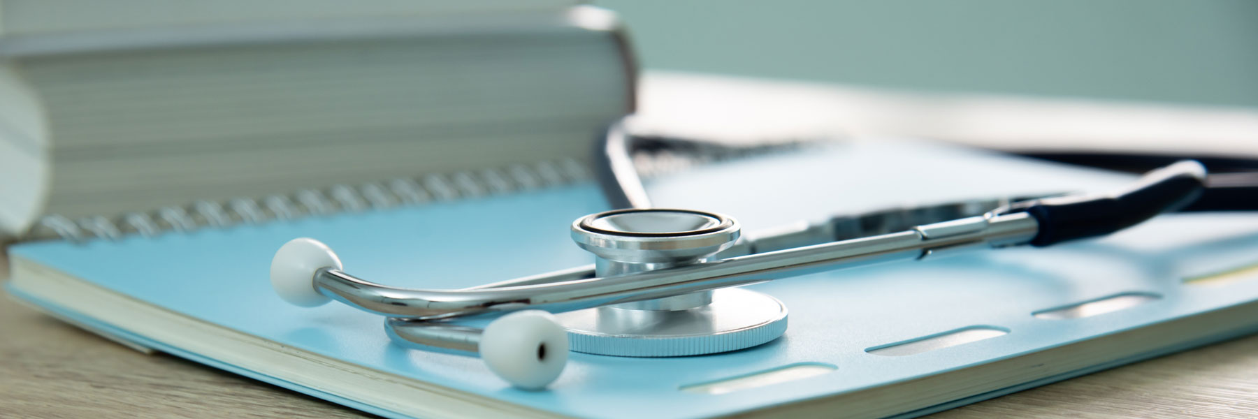 Image of a notebook with stethoscope on top of it, as well as other textbooks in the background, all setting on a table